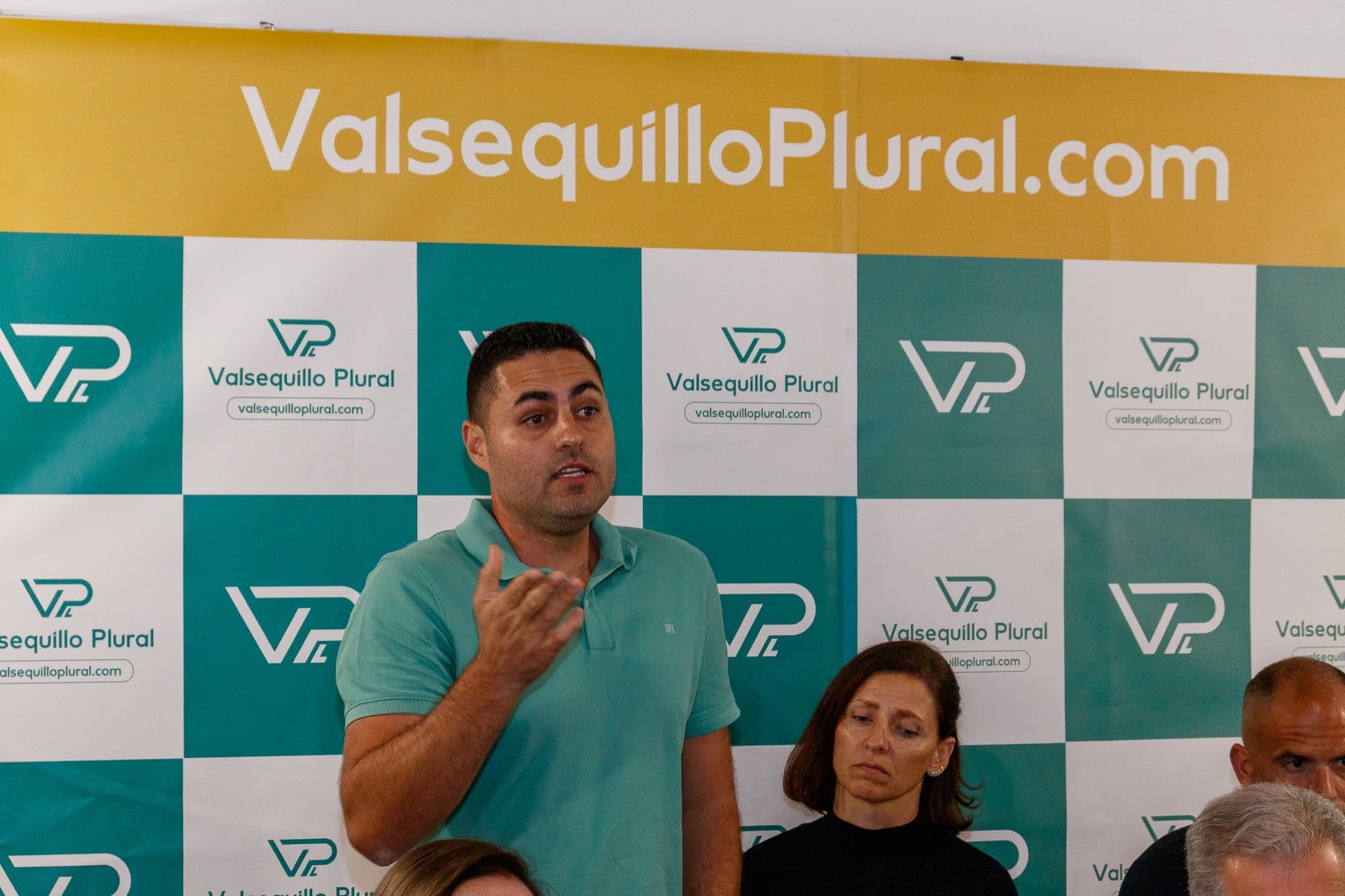Un hombre con polo verde habla delante de un panel repetido con el logotipo de Valsequillo Plural y la dirección valsequilloplural.com.