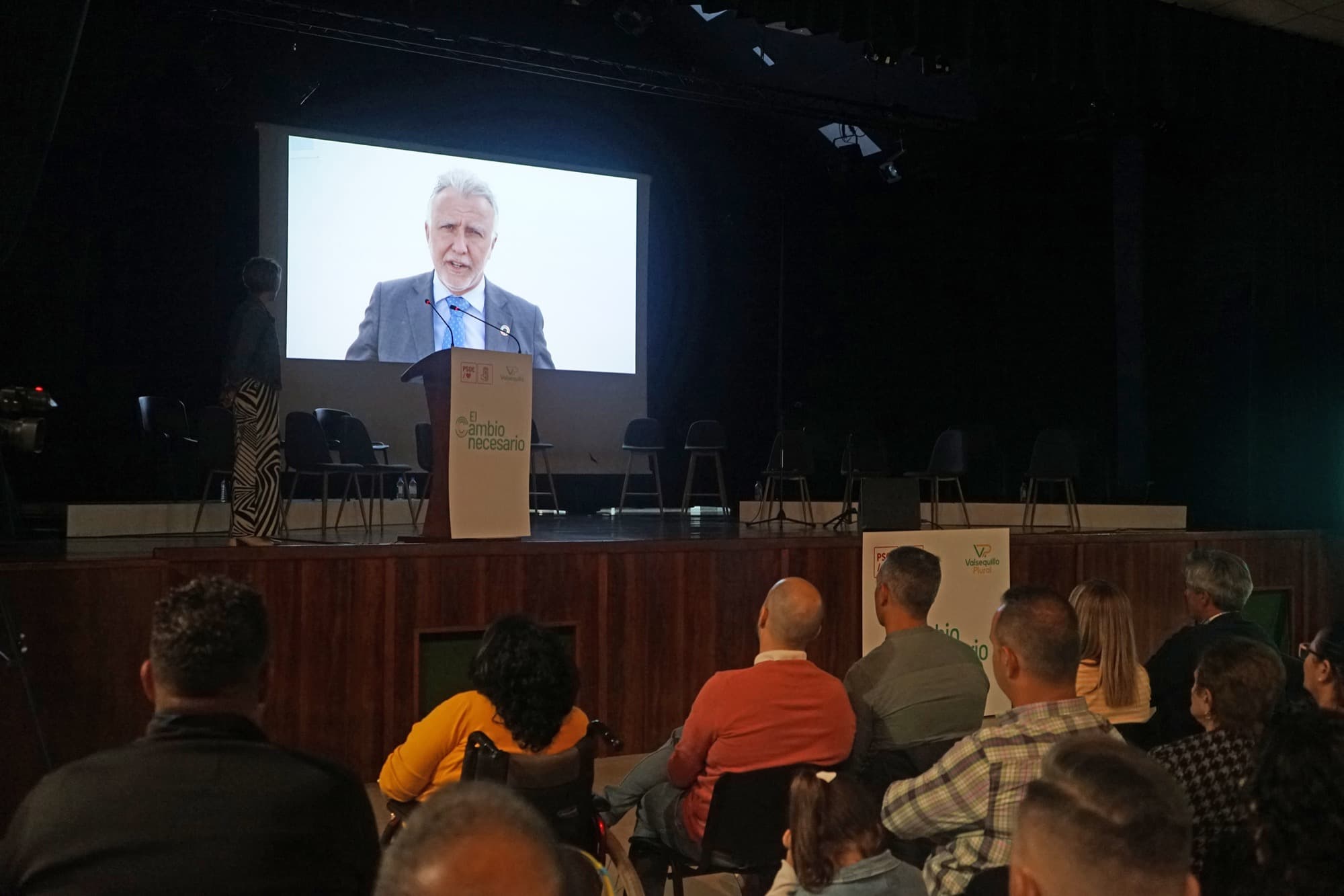 Un ponente interviene desde un atril mientras su imagen se proyecta ampliada en la pantalla, ante el público sentado.