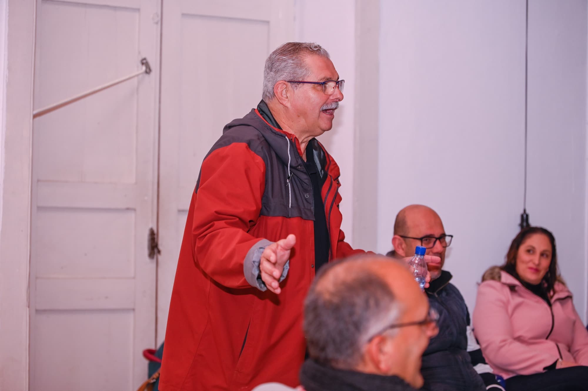 Un hombre con chaqueta roja, de pie, habla y gesticula ante un público sentado en una sala.