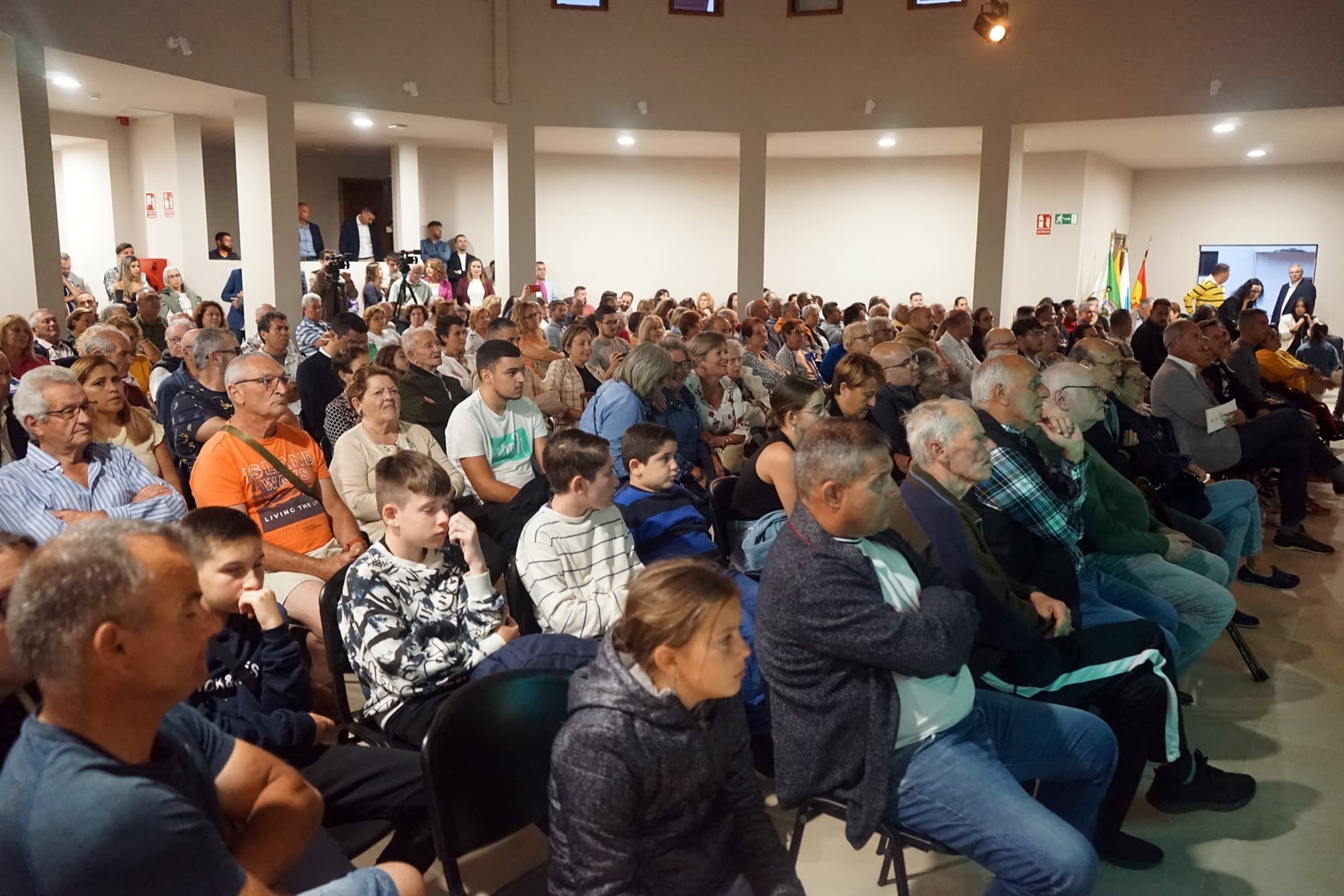 Sala llena de gente de todas las edades, sentada y escuchando un acto público.